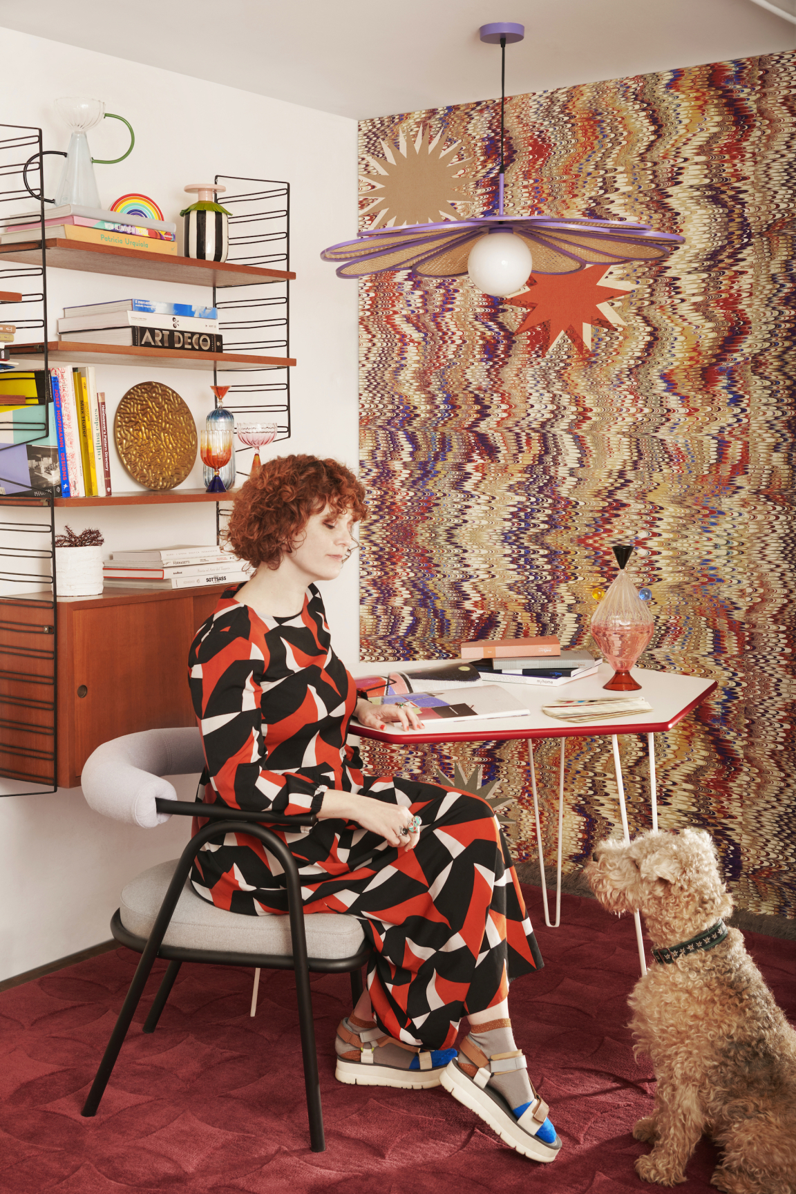 A woman in a patterned dress sits at a modern desk, accompanied by a fluffy dog in a stylish interior.