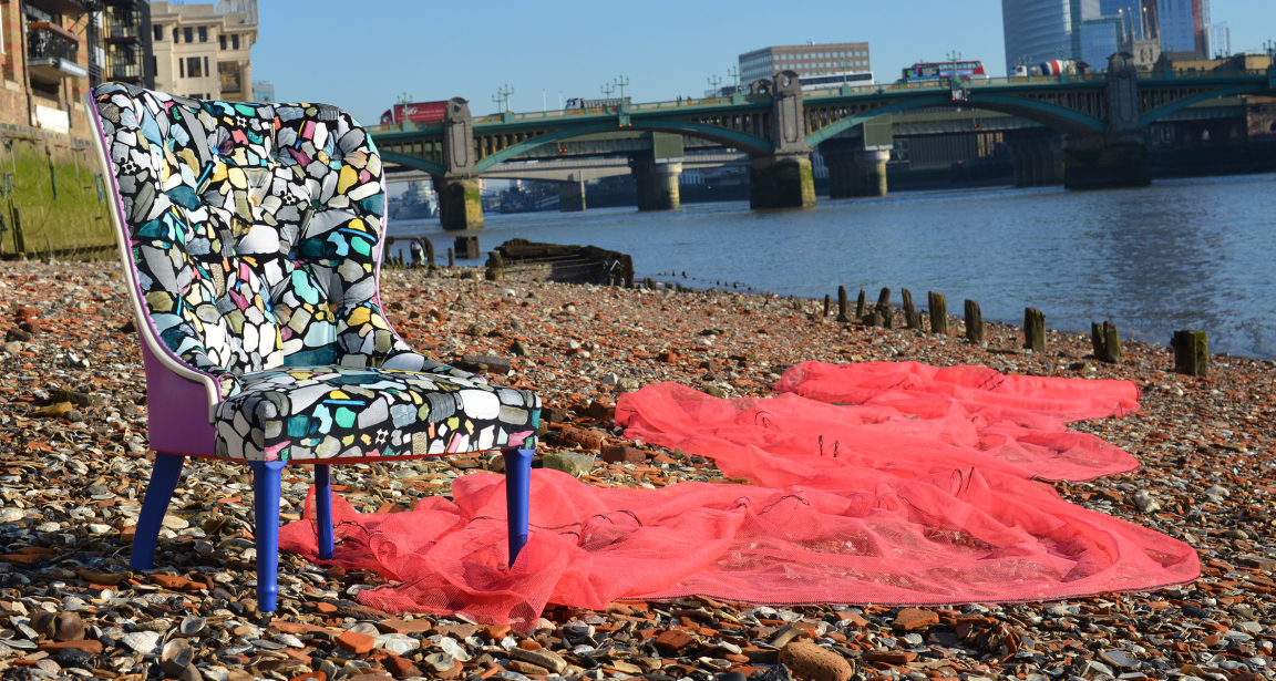 Colorful, abstract-patterned chair on a pebble beach, with pink fabric draped nearby and a bridge in the background.