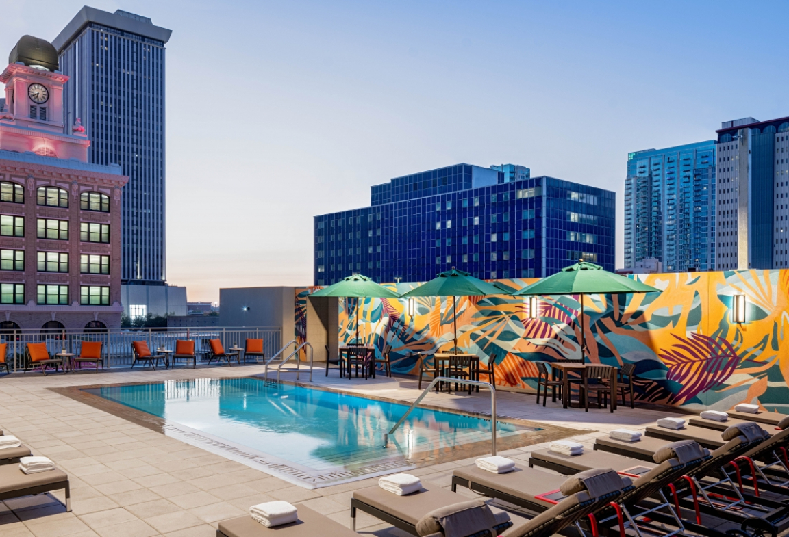 Rooftop pool area featuring vibrant murals, lounge chairs, and umbrellas against a city skyline at twilight.