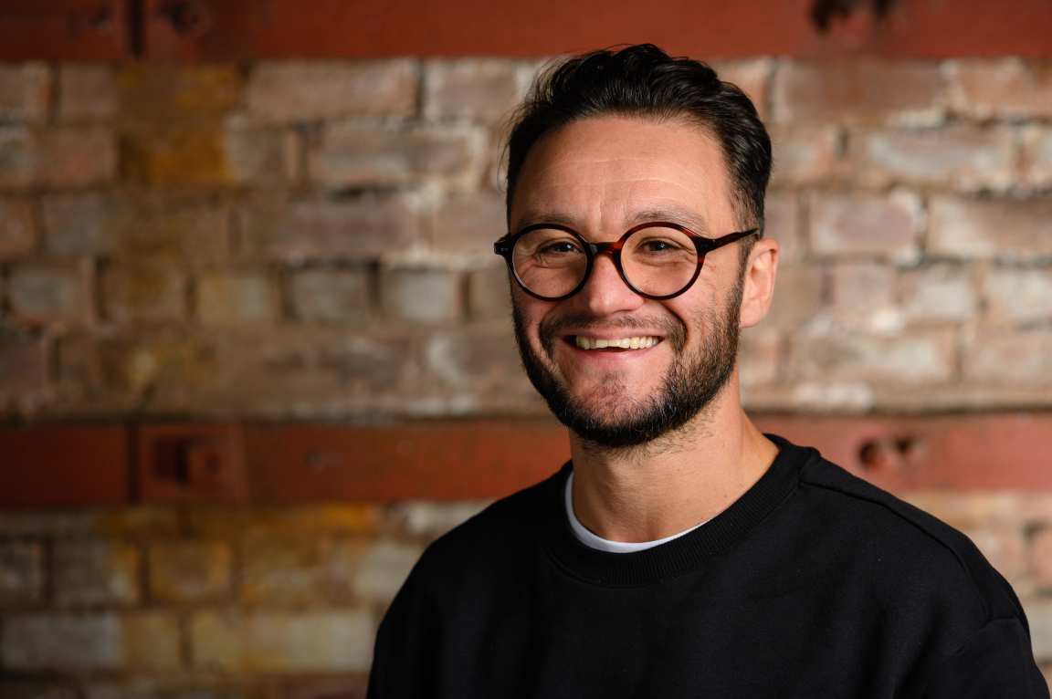 Andrew Hadden, HLM Architects associate, smiles against a rustic brick backdrop, highlighting sustainable architecture and carbon reduction themes.