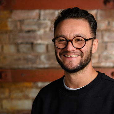 Andrew Hadden, HLM Architects associate, smiles against a rustic brick backdrop, highlighting sustainable architecture and carbon reduction themes.