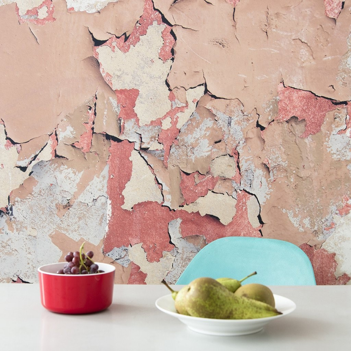 Textured wall with peeling paint in warm tones, featuring red and beige colors, alongside a bowl of grapes and pears.