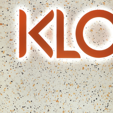 Illuminated orange "KLO" logo on a textured, speckled wall designed for a travel technology office.