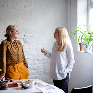 Rachel Withey and Sarah Dodsworth engaged in conversation at Ekho Studio, surrounded by artistic materials and plants.