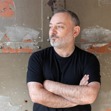 Creative director Jon Humphreys in a black shirt, showcasing a thoughtful expression against a textured industrial backdrop.