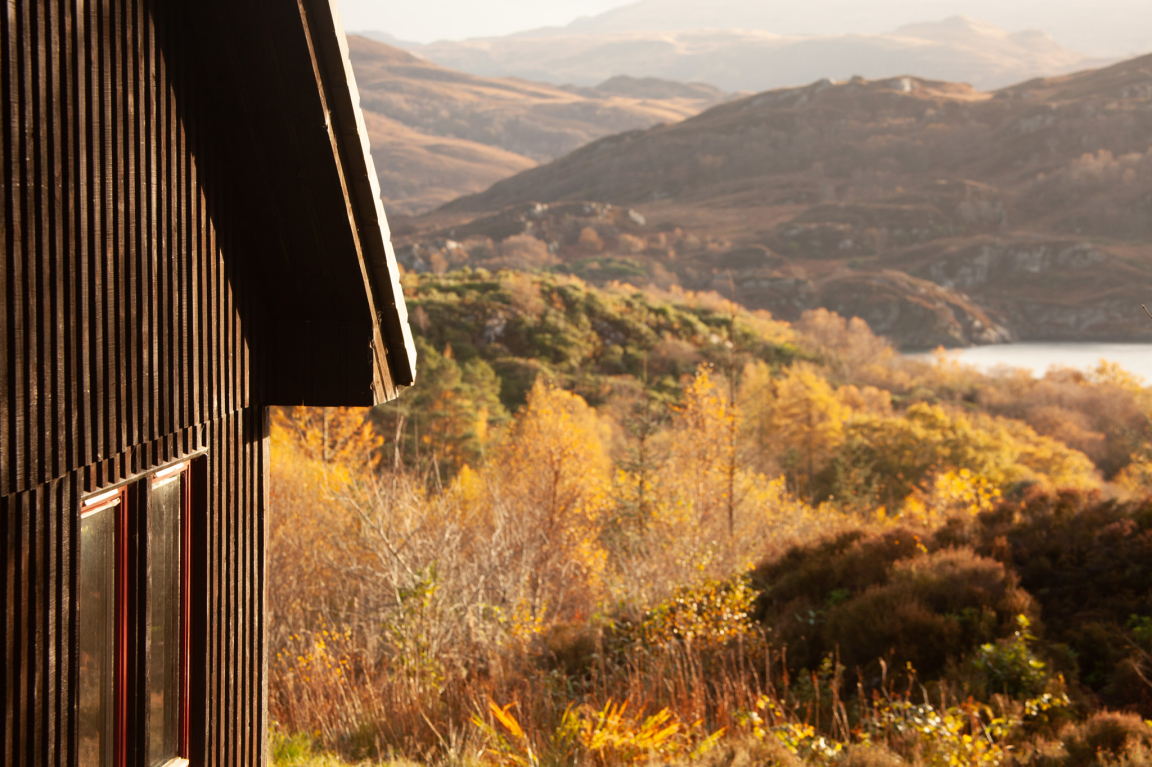 Black timber cabin with large windows nestled in a vibrant autumn landscape, showcasing Hebridean architectural influence.